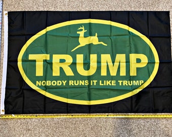 May include: A black flag with a green and yellow oval. The oval has a silhouette of a deer and the text "TRUMP" in yellow letters. Below the oval is the text "NOBODY RUNS IT LIKE TRUMP".