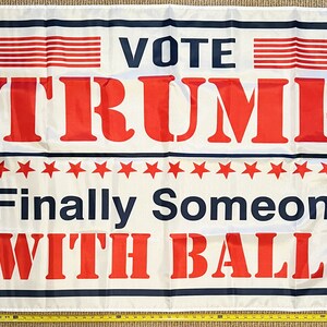 May include: White flag with blue and red text that reads "Vote Trump Finally Someone With Balls". There are 16 red stars and a blue border around the text.
