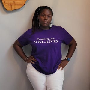 May include: A woman wearing a purple t-shirt with the text "No makeup, just MELANIN" in white. She is wearing white shorts and has her hair in dreadlocks.