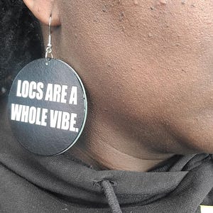 May include: Black circular wooden earrings with the text "LOCS ARE A WHOLE VIBE." in white lettering.