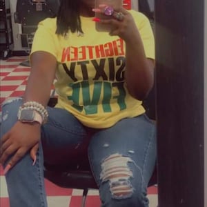 May include: A person wearing a yellow t-shirt with the text "Sixteen Nineteen Eighty Five" in red, green, and black. They are wearing blue jeans with rips in the knees and a brown baseball cap with a black fist symbol.