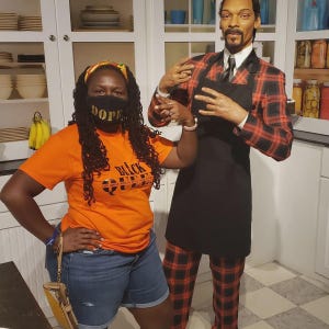 May include: A woman wearing a black face mask with the word "DOPE" on it and an orange t-shirt with the text "BLACK QUEEN" stands next to a wax figure of Snoop Dogg wearing a red and black plaid suit and a black apron.