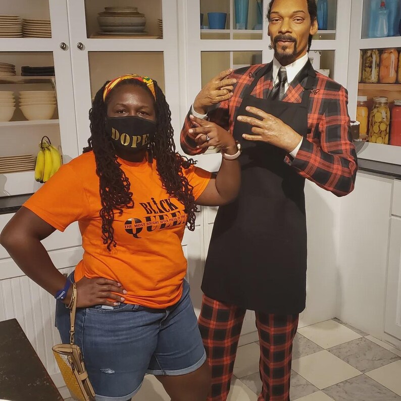 May include: A woman wearing a black face mask with the word "DOPE" on it and an orange t-shirt with the text "BLACK QUEEN" stands next to a wax figure of Snoop Dogg wearing a red and black plaid suit and a black apron.