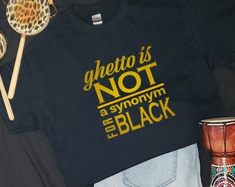 May include: Black t-shirt with the text "ghetto is NOT a synonym FOR BLACK" in gold lettering. The shirt is paired with light blue jeans and a small drum. A pair of percussion instruments with animal print heads are also visible.