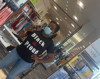 May include: A person wearing a black t-shirt with the text "Black & Proud" in white lettering and a red heart symbol. The person is wearing blue jeans and a blue face mask.