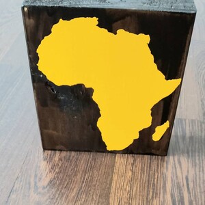 May include: A wooden block with a yellow silhouette of the continent of Africa painted on it. The block is stained dark brown.