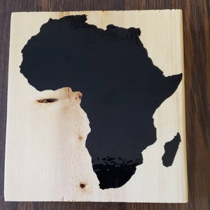 May include: A wooden plaque with a black silhouette of the continent of Africa. The plaque is made of light wood with a natural grain.