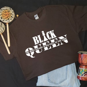 May include: A brown t-shirt with a white graphic design that says "Black Queen" with a chess piece graphic and the text "The most sought after piece in the game."