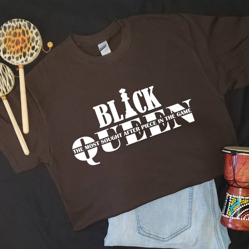 May include: A brown t-shirt with a white graphic design that says "Black Queen" with a chess piece graphic and the text "The most sought after piece in the game."