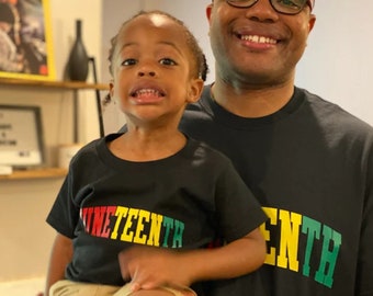 May include: A father and son wearing black t-shirts with the word "NINETEENTH" printed in red, yellow, and green. The son is sitting on the father's lap.