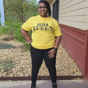 May include: A woman wearing a yellow t-shirt with the text "Black Queen The Most Sought After Piece In The Game" in black lettering. She is wearing black pants and black sneakers.