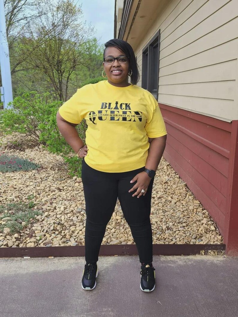 May include: A woman wearing a yellow t-shirt with the text "Black Queen The Most Sought After Piece In The Game" in black lettering. She is wearing black pants and black sneakers.