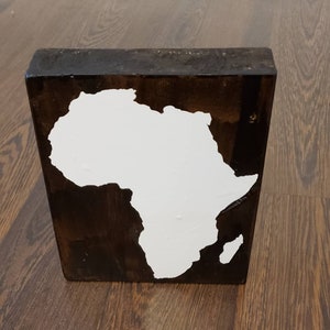 May include: A wooden block with a white painted outline of the continent of Africa. The block is dark brown and has a rustic finish.