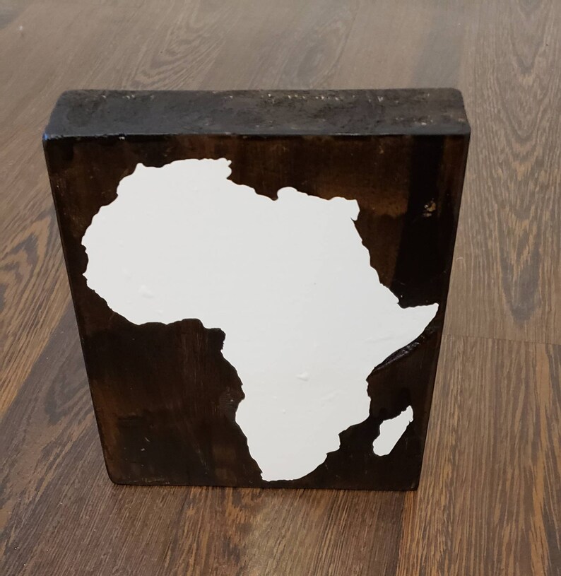 May include: A wooden block with a white painted outline of the continent of Africa. The block is dark brown and has a rustic finish.