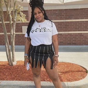 May include: A woman wearing a white t-shirt with the text "Black Queen" printed on it. She is also wearing a black skirt with zipper details and a silver watch.
