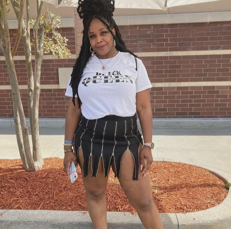 May include: A woman wearing a white t-shirt with the text "Black Queen" printed on it. She is also wearing a black skirt with zipper details and a silver watch.