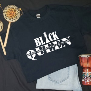 May include: Black t-shirt with white text that reads "Black Queen The Most Sought After Piece In The Game". The text is stylized to look like a chess piece.