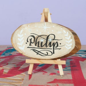 May include: A wooden sign with the name "Philip" written in black calligraphy on a white background. The sign is decorated with white snowflakes and leaves and is displayed on a small wooden easel.