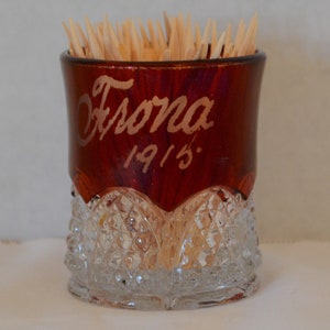 May include: A red glass toothpick holder with a clear glass base. The holder has the word "Escona" and the year "1915" written on it.