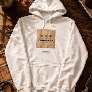 Op de afbeelding: Een witte hoodie met een beige rechthoekige patch op de borst met de tekst BURBERRY London England en drie cirkelvormige grafische iconen erboven. Een kleiner wit rechthoekig label met de tekst BURBERRY bevindt zich daaronder.