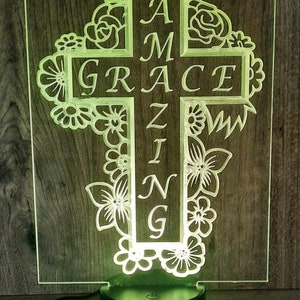 May include: A white cross with floral designs around it. The cross has the words "AMAZING GRACE" written on it. The cross is illuminated with a green light.