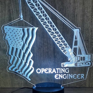 May include: A lighted acrylic sign featuring a crane and an American flag design. The words "OPERATING ENGINEER" are displayed below the crane. The sign is mounted on a black base and is illuminated with a bright white light.