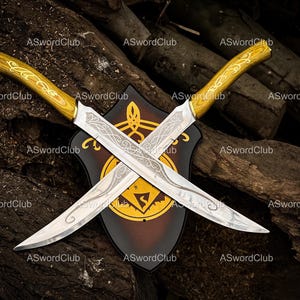 May include: Two crossed silver daggers with etched blades and wooden handles. They are displayed against a shield with a gold emblem and a dark brown background. The handles have a gold accent.