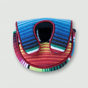 Puede incluir: Una colorida funda para putter de golf con un diseño de serape a rayas. La funda presenta una vibrante mezcla de rayas azules, rojas, amarillas y verdes, delineadas con ribete rojo. Tiene un interior negro y una abertura en forma de U.