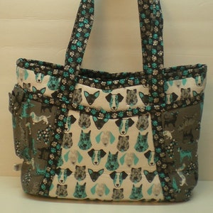 May include: A black and teal tote bag with a pattern of dog faces and paw prints. The bag has two straps and a zippered pocket on the front.