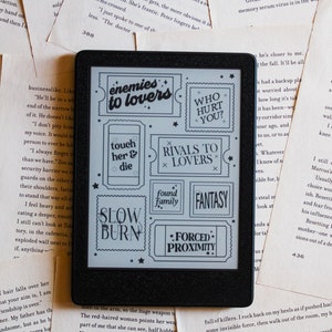 May include: A black and white e-reader with a screen displaying a variety of book genre labels, including "enemies to lovers", "touch her & die", "slow burn", "forced proximity", "who hurt you?", "rivals to lovers", "found family", and "fantasy".