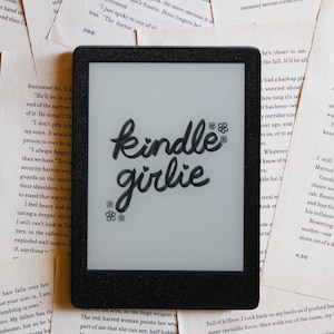 May include: A black and white Kindle e-reader with the words "kindle girlie" written in black cursive on the screen.