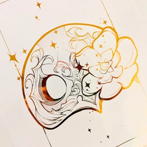 May include: A gold foil print on white paper featuring a decorative mask design with floral and star accents. The mask has a crescent moon shape and intricate detailing. The artwork is set against a white background with a gold border.