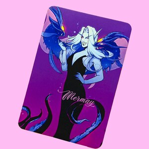 May include: A purple card with a woman with long blue hair and a black dress. She is holding two blue creatures with yellow eyes. The card says "Mermay" in pink cursive.