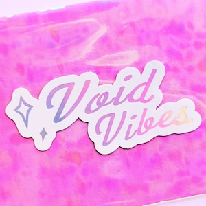 May include: A holographic sticker with the text "Void Vibes" in a white cursive font with a rainbow gradient. The sticker has a small star design on the left side.