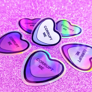 May include: Five iridescent holographic heart-shaped stickers with the text "Corrupt Me" on each. The stickers are on a pink sparkly background.