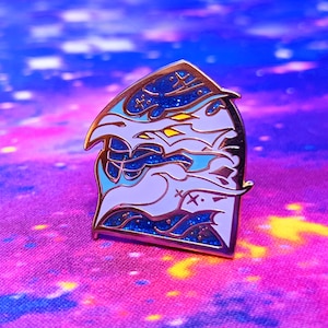 May include: A white and blue enamel pin with a glittery finish. The pin features a stylised design of a creature with a large head and a long, flowing tail. The pin is shaped like a house.