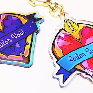 May include: Two heart-shaped acrylic charms with gold keychains. The first charm features a purple and blue heart with the text "Sailor Void". The second charm features a red and pink heart with the text "Sailor Sunwell".