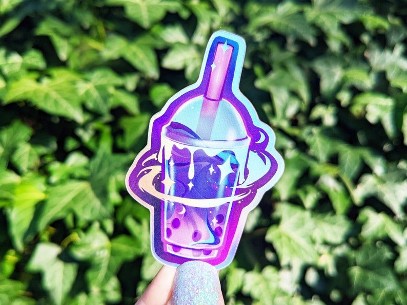 May include: A holographic sticker of a galaxy-themed boba tea with a straw and boba pearls. The sticker is iridescent and has a purple and blue color scheme.