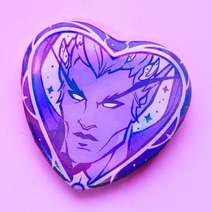 May include: A heart-shaped badge featuring a purple and blue illustration of a male figure with pointed ears and a crown. The badge has a white border and is set against a pink background.