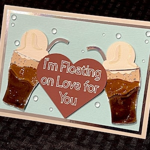 May include: A handmade greeting card with a light blue background and two root beer floats. A brown heart in the center reads "I'm Floating on Love for You". The card has a white border and silver accents.