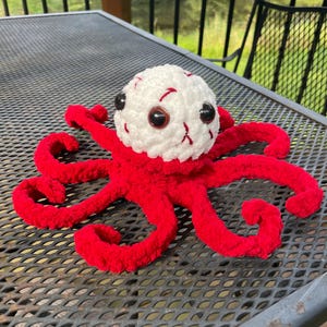 May include: A handmade, plush octopus toy with a white head and red tentacles. The head features large, black eyes and red detailing. The octopus is sitting on a metal surface, with a natural outdoor background.