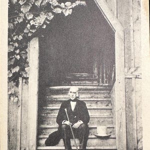 May include: A black and white photograph of a man sitting on steps in front of a wooden building. The man is wearing a suit and holding a cane. The text below the image reads "MR. ALCOTT IN THE DOOR OF THE SCHOOL OF PHILOSOPHY Orchard House Concord, Massachusetts."