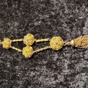 May include: A gold chain necklace with a tassel and three ornate round charms. The charms are detailed with intricate designs.