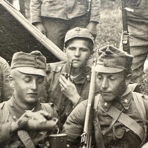 May include: Black and white photograph of a group of soldiers in uniform, wearing caps and holding rifles. One soldier is smoking a pipe. The image appears to be from a historical event.