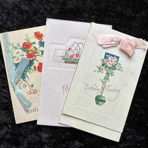 May include: Three vintage birthday greeting cards. One card features a floral design with red poppies and blue ribbon. Another has a floral illustration with the text "Birthday Greeting". The third card has a minimalist floral design.