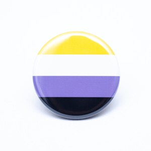 May include: A round button with the colors of the non-binary pride flag. The button is yellow, white, purple, and black.
