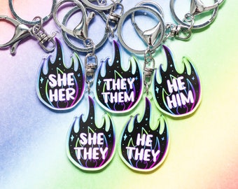 Può includere: Cinque portachiavi colorati a forma di fiamma con il testo "She Her", "They Them", "He Him", "She They" e "He They" su ogni portachiavi.