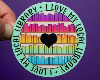May include: A round sticker with the text "I LOVE MY LOCAL LIBRARY" in a circular pattern. The sticker features a rainbow-colored bookshelf design with a black heart in the center. The background is a solid purple.