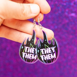 May include: A pair of black and white flame-shaped earrings with the text "They Them" on each earring. The earrings are made of clear acrylic and have silver hooks.