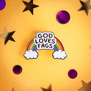 May include: A rainbow enamel pin with the text "GOD LOVES FAGS" in black letters.